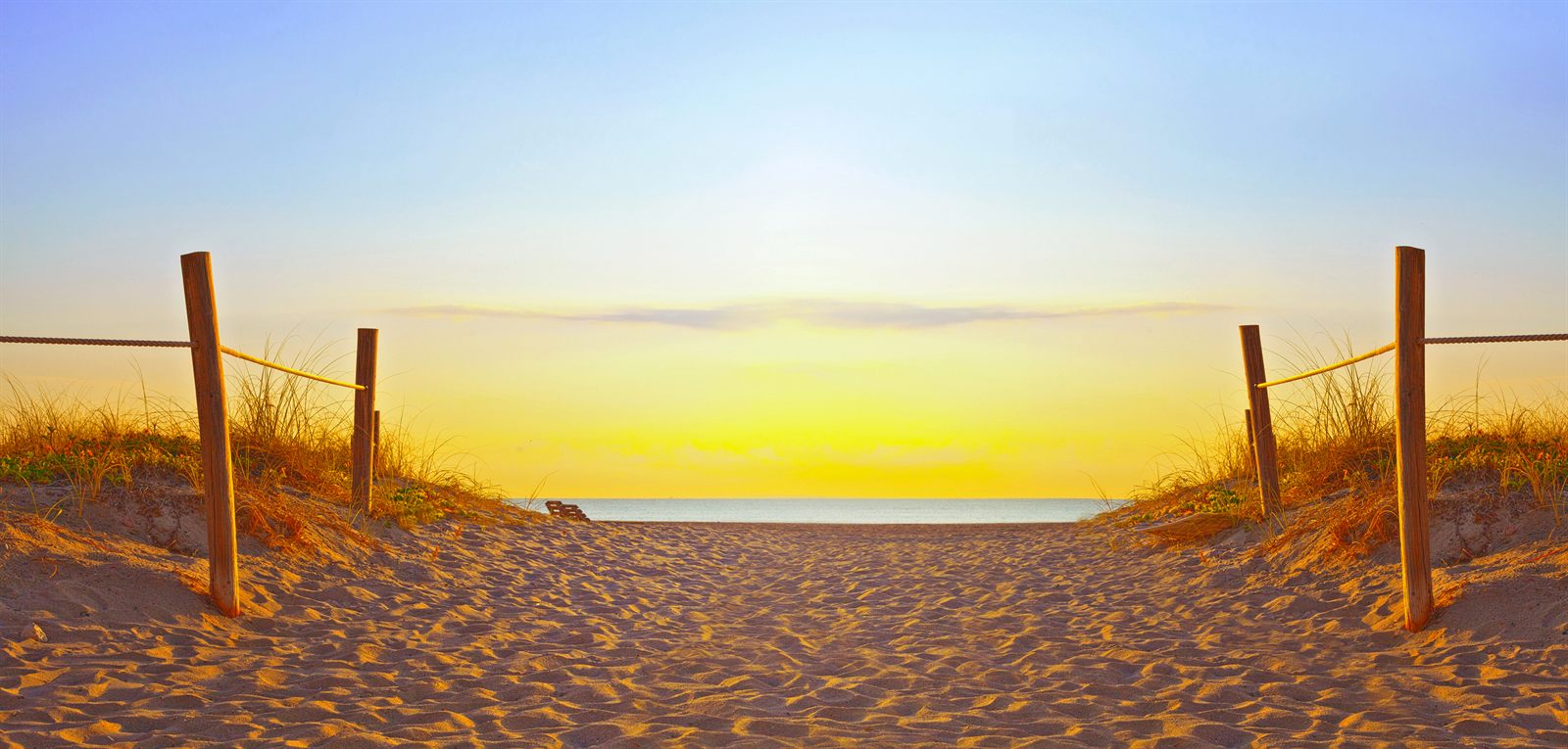 A sandy beach path between rope fences and dune grass leading to the ocean at sunrise